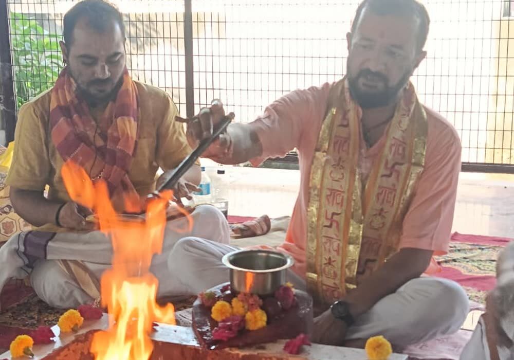 Two Brahmins seated at a square fire altar in Vrindavan, offering ghee into the flames, the rim of the kund laid with marigolds and roses.